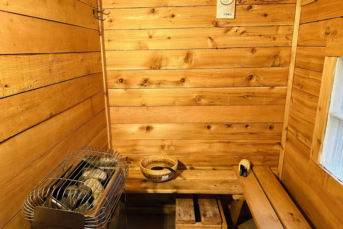 A warm, inviting sauna interior lined with natural wooden walls, showcasing a traditional sauna stove, a wooden bench, and a bowl for water or essential oils. The scene exudes serenity, with subtle hints of wellness rituals and shared moments.