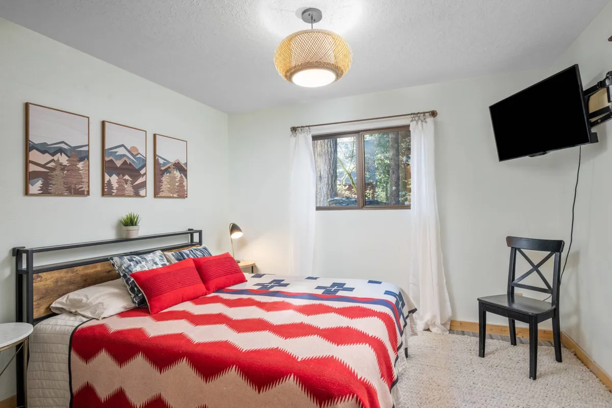 A queen-sized bed with a red and white patterned blanket is the centerpiece, accompanied by mountain-themed artwork above, a window with sheer white curtains letting in natural light, and a black chair in the corner. The atmosphere is cozy and peaceful, ideal for unwinding.