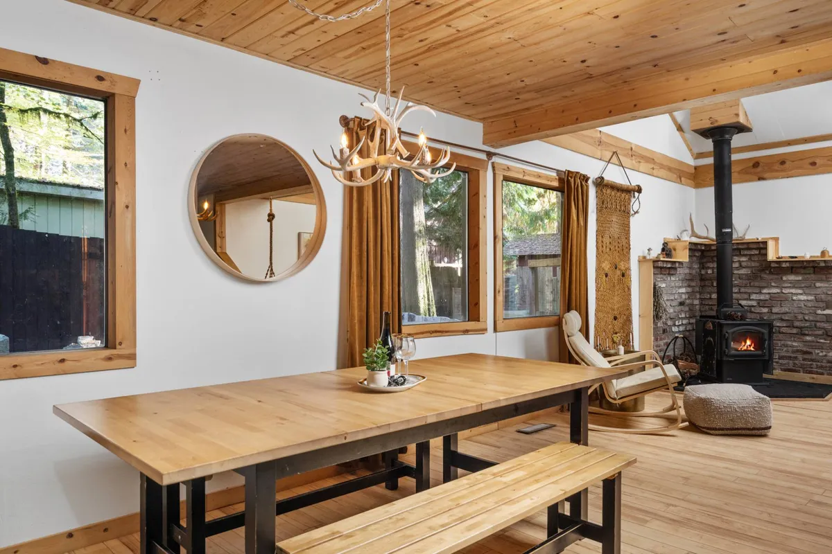 This photo showcases a wooden dining table set near a fireplace and large windows, with a chandelier illuminating the space. The arrangement suggests a welcoming spot for group meals, game nights, or morning coffee, all while enjoying views of the outdoors.