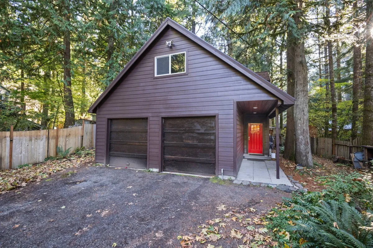 The exterior view highlights the red front door, wooden siding, garage doors, and abundant surrounding trees. The driveway is visible, offering space for parking and easy access to outdoor adventures.