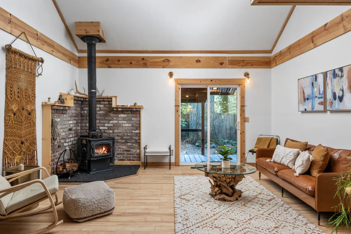 This image captures a warm, open living area featuring a leather sofa, decorative rug, and a wood-burning stove. Sliding glass doors fill the room with natural light and provide glimpses of the surrounding forest, while the layout invites relaxed gatherings and cozy moments by the fire.