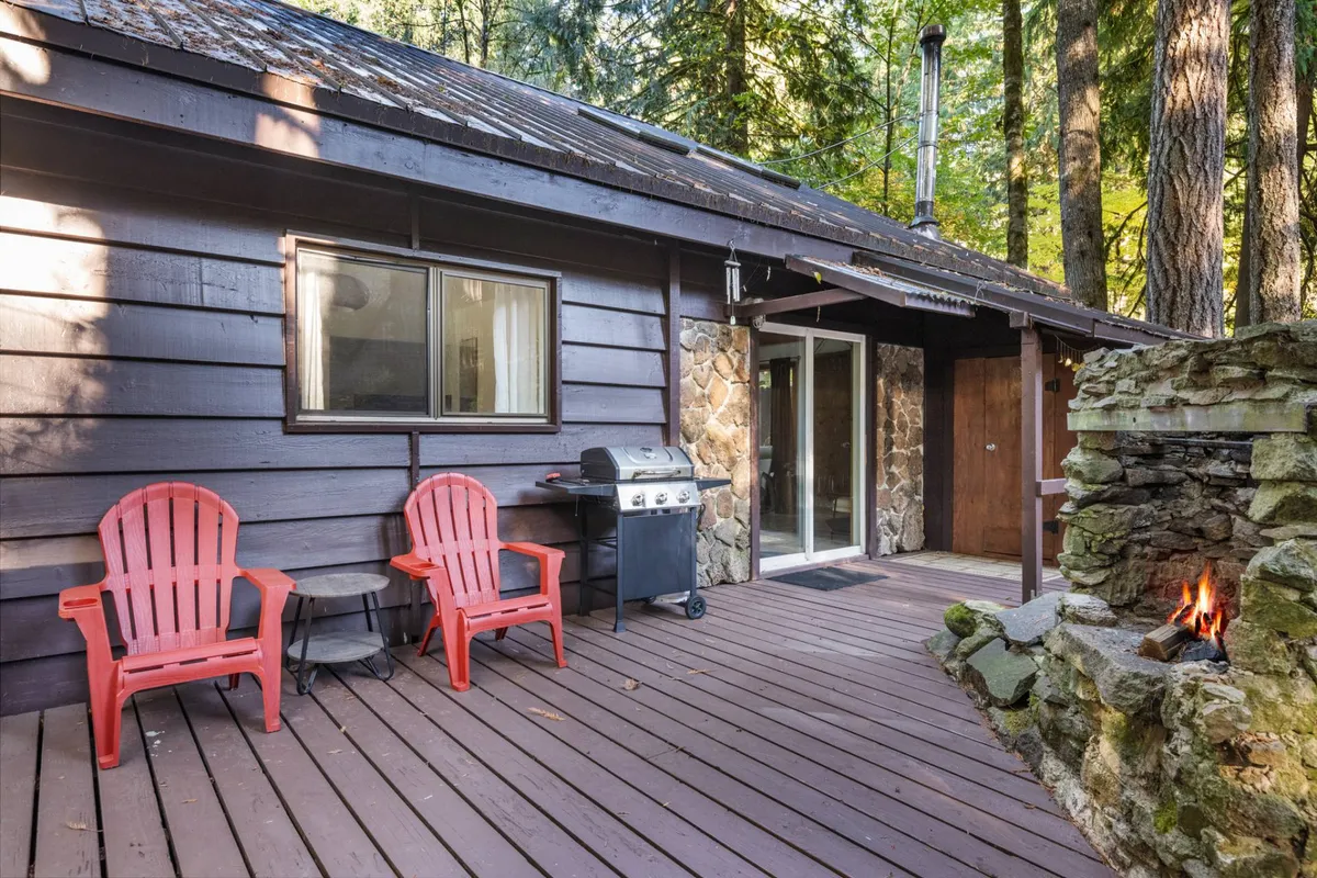 This exterior image showcases a deck with red Adirondack chairs, a stone fireplace, and a grill, set against a backdrop of lush trees. The scene invites relaxation and socializing, with plenty of space to unwind after outdoor adventures.