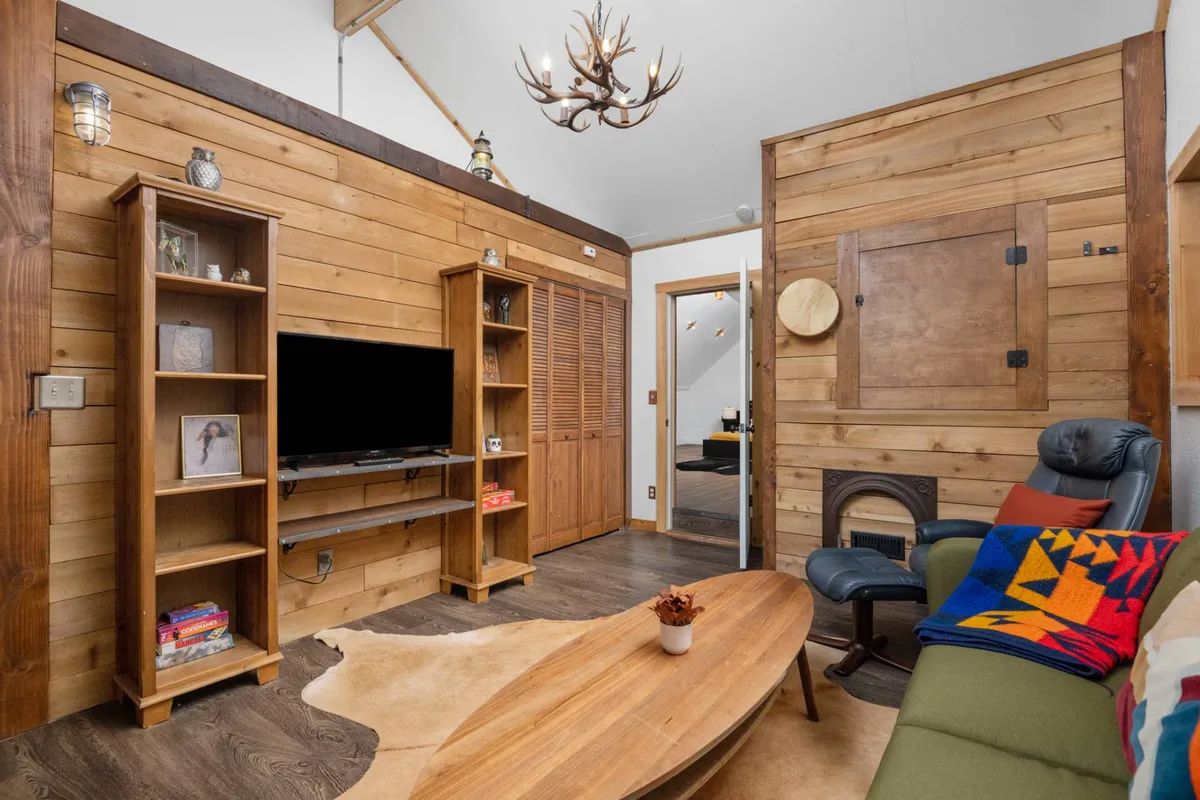 A welcoming loft-style area with wooden shelves, a TV unit, round coffee table, a colorful throw blanket, and an antler chandelier. The setting is ideal for socializing, watching TV, or relaxing, with warm lighting and inviting decor.