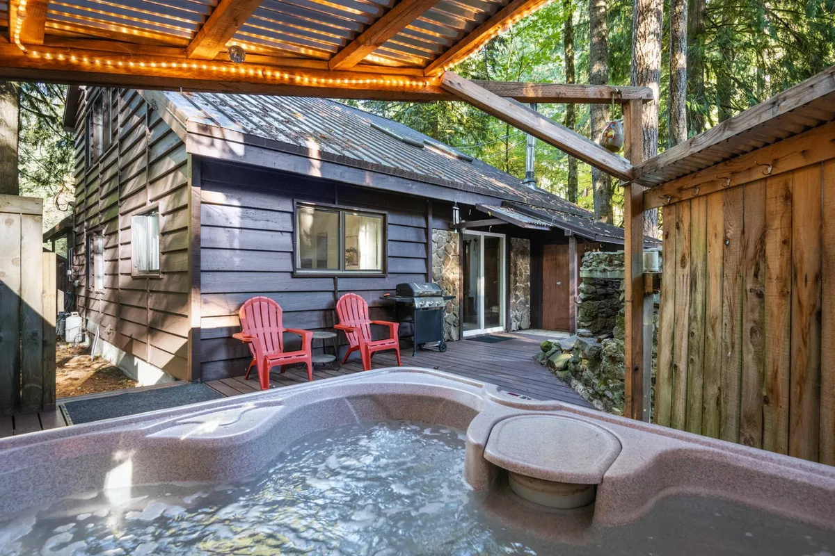 A cozy outdoor space featuring a bubbling hot tub under a wooden pergola, surrounded by red chairs and a view of the home's exterior. The scene suggests warmth and relaxation even on snowy days, with hints of outdoor enjoyment like barbecuing or stargazing.
