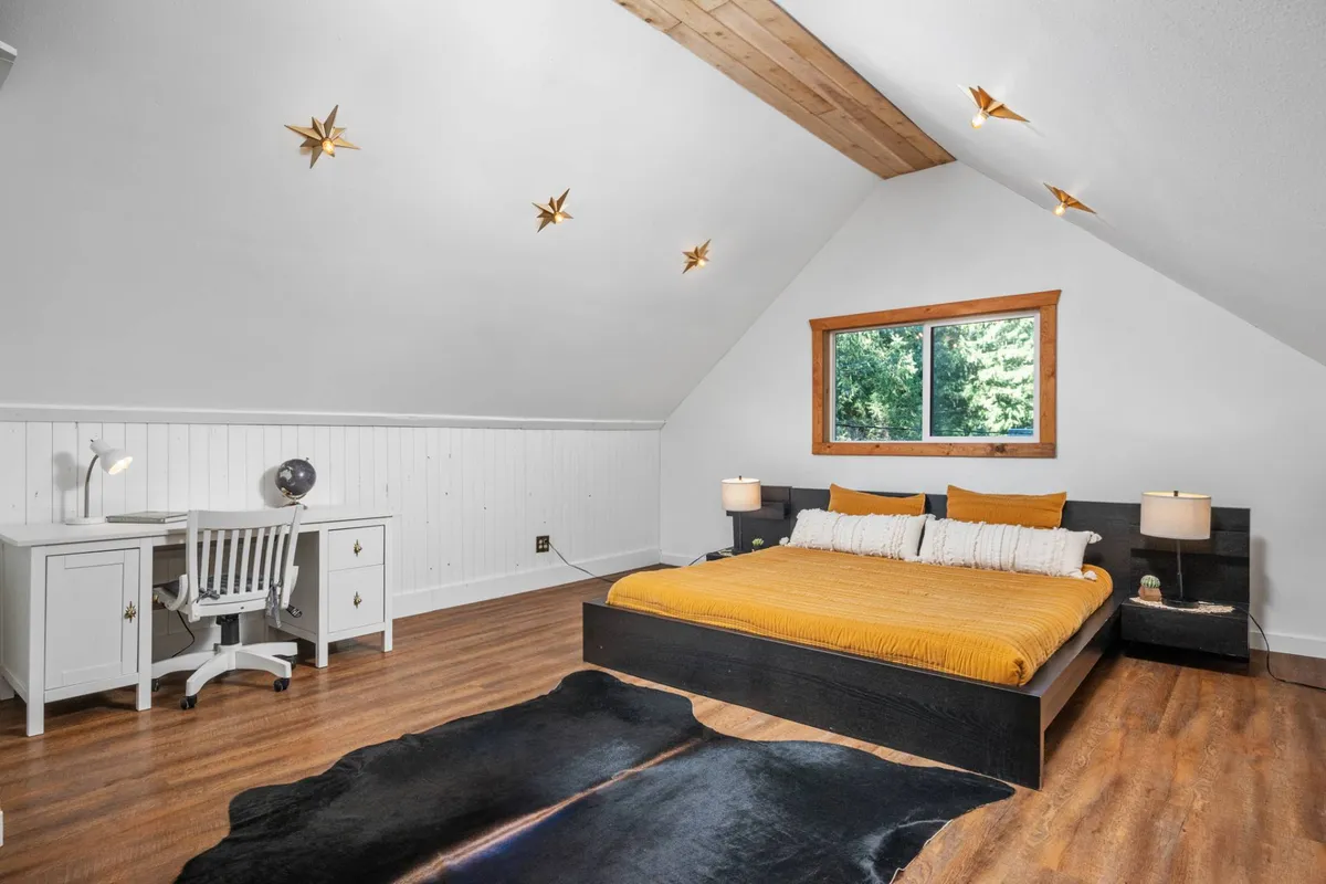 A bed with mustard yellow bedding stands next to a home office desk, set atop a cowhide rug. A large window frames a forest view, infusing the room with natural light. The combination of sleeping and work space suggests versatility and comfort.