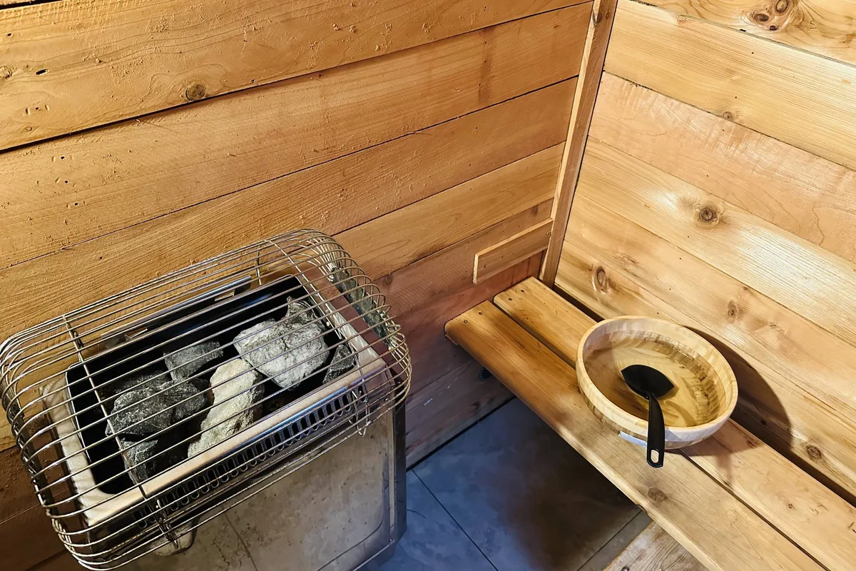 A tranquil interior sauna space with a wooden bench, heating unit, and a wooden bowl with ladle, inviting guests to relax and unwind. The setting is intimate and promises rejuvenation after outdoor winter adventures.