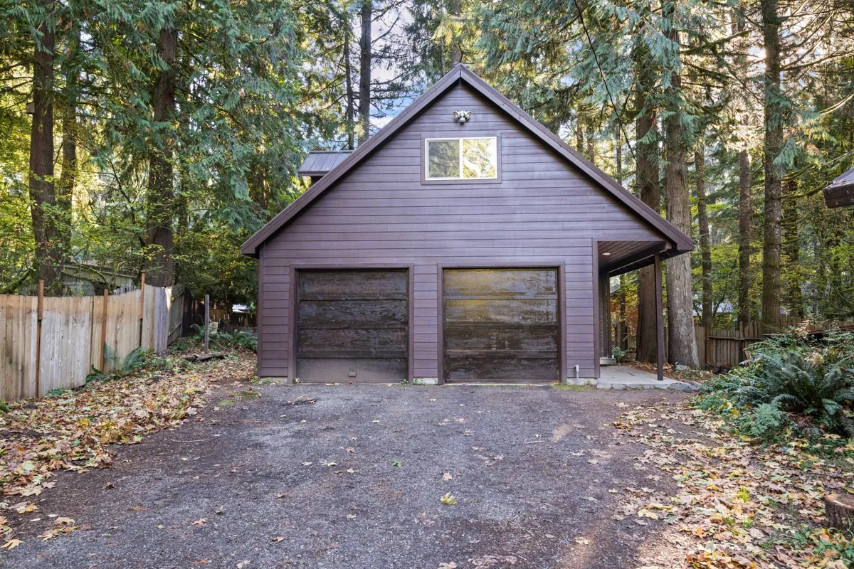 This exterior image showcases two garage doors on the wooden house, surrounded by tall trees, and a spacious area in front suitable for parking vehicles, RVs, or trailers. The setting is inviting and hints at ample storage or parking flexibility.