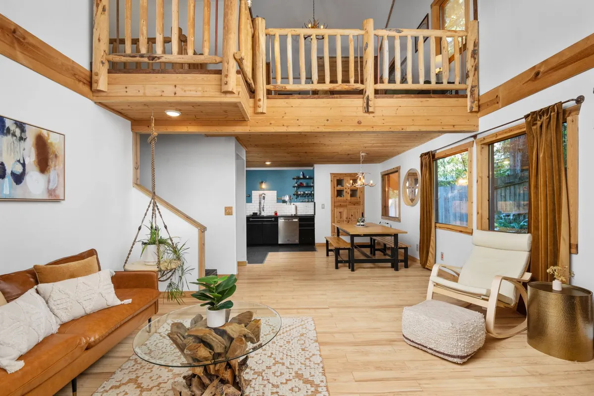This inviting living area showcases rustic wooden beams, a plush leather sofa, and a dining table set beside expansive windows that look out onto lush forest greenery, all bathed in the soft glow of a hanging light fixture. The scene radiates warmth and comfort, perfect for gathering with loved ones or unwinding after a day in the mountains.
